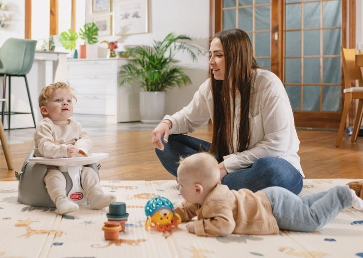 A woman on a Nuby playmat with two young children. One child is crawling toward Nuby toys. The other is sitting in a Nuby My Floor Seat with Activity Tray.