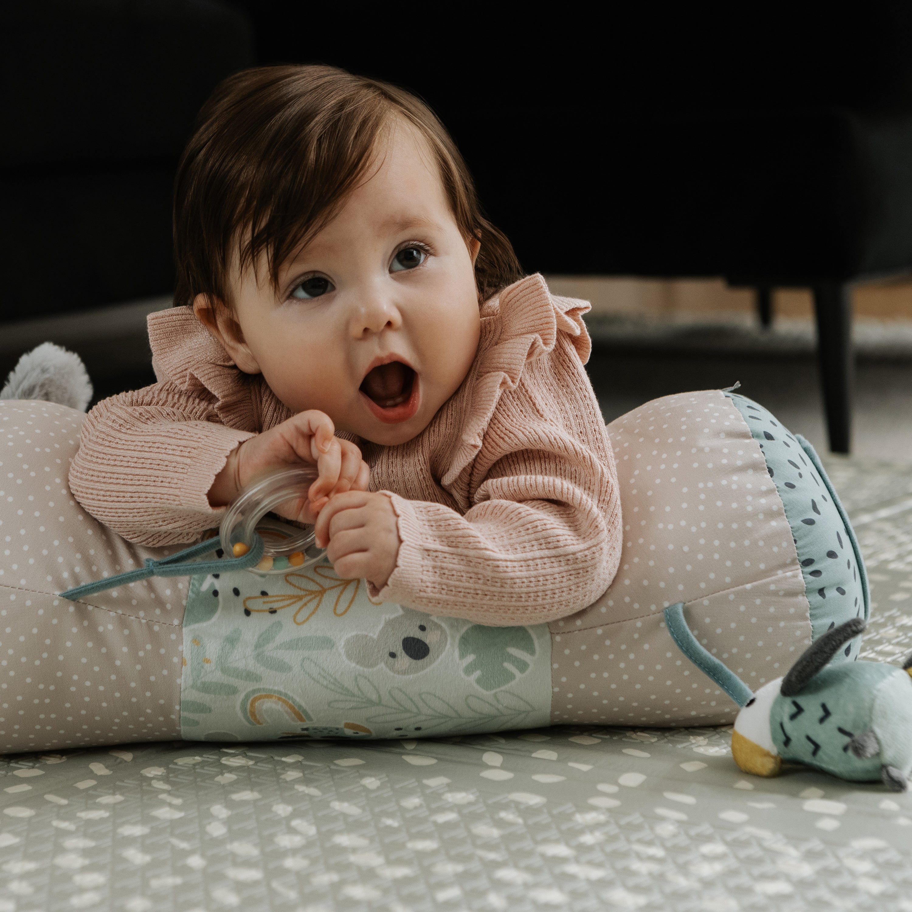 A baby with brown hair in a pink sweater lies on a patterned mat, propped up on the Animal Adventures Tummy Time Pillow with an attached stuffed toy, holding a teething ring and gazing upward with an open mouth.
