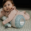 A baby in a pink outfit lies on a patterned mat, hugging the Animal Adventures Tummy Time Pillow—a sensory baby pillow with a mirror and animal design—while looking up with a gentle expression.