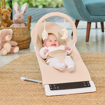 A baby in white smiles in the Deluxe Baby Bouncer Seat | Khaki with hanging toys. Plush animals rest nearby, enhancing the cozy, sunlit room’s charm.