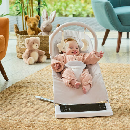 A smiling baby sits in the Deluxe Baby Bouncer Seat | Beige, enjoying hanging toys. Plush teddy bears and a bunny rest on a woven rug in a cozy, sunlit room with colorful chairs.