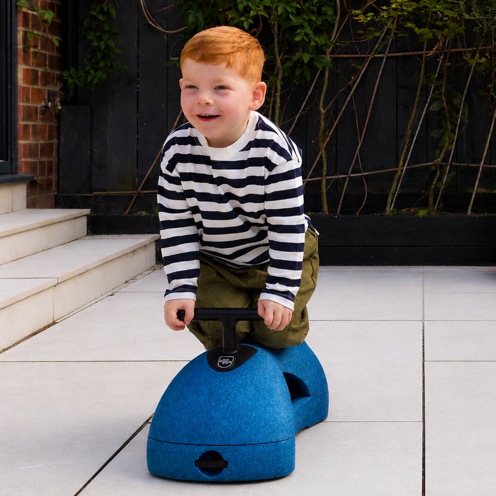A young child with red hair, wearing a striped shirt and green pants, kneels on the Boogie Buggy | Blue outdoors on a tiled patio.