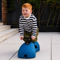 A young child with red hair, wearing a striped shirt and green pants, kneels on the Boogie Buggy | Blue outdoors on a tiled patio.