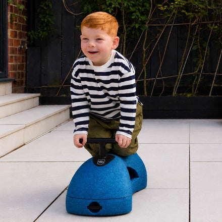 A young child with red hair, wearing a striped shirt and green pants, kneels on the Boogie Buggy | Blue outdoors on a tiled patio.
