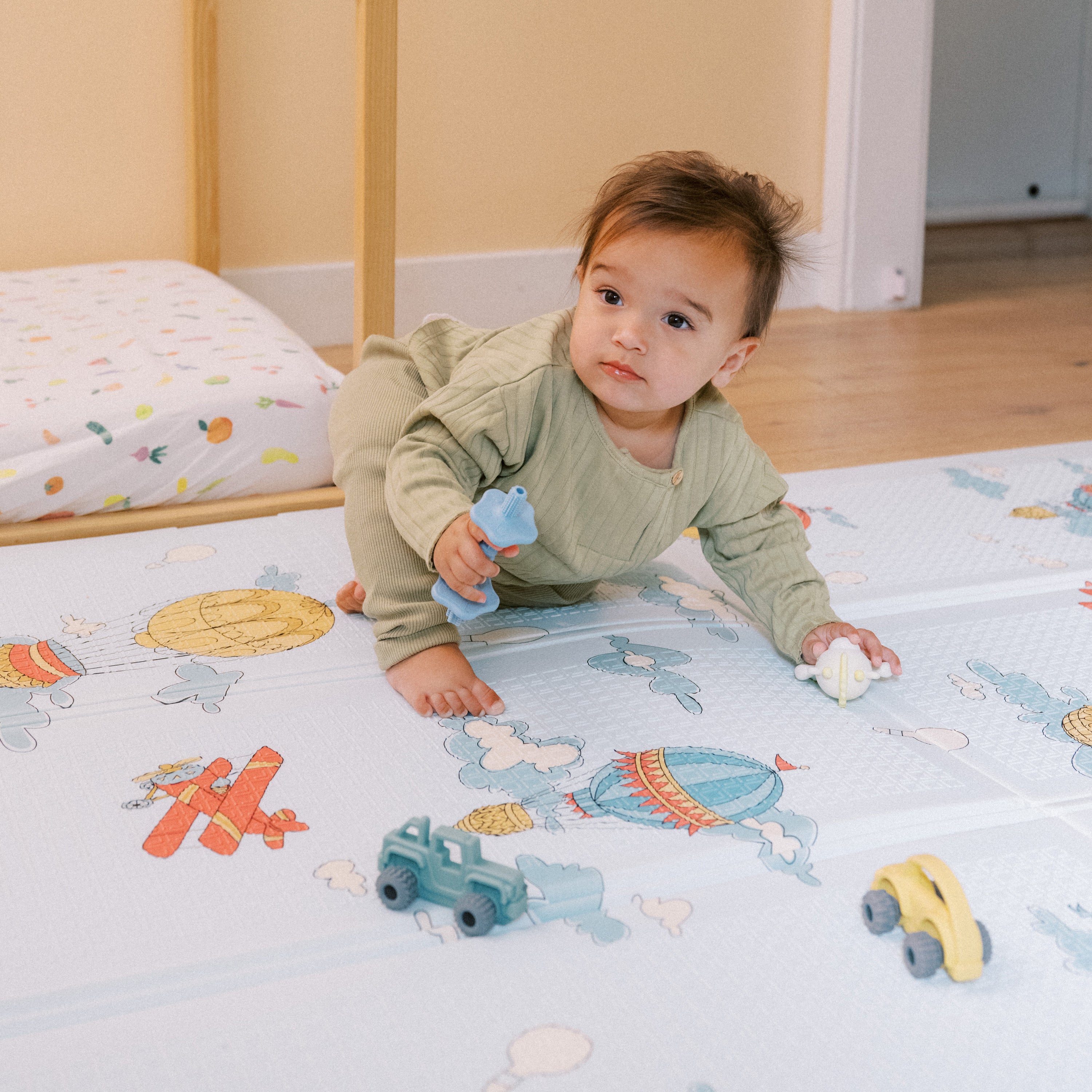 A baby in green crawls on the Reversible Floor Mat | Hot Air Balloons, which features colorful hot air balloon and airplane designs, while holding a blue toy and white ball. Toy vehicles are scattered around the portable play mat.