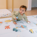 A baby in green crawls on the Reversible Floor Mat | Hot Air Balloons, which features colorful hot air balloon and airplane designs, while holding a blue toy and white ball. Toy vehicles are scattered around the portable play mat.