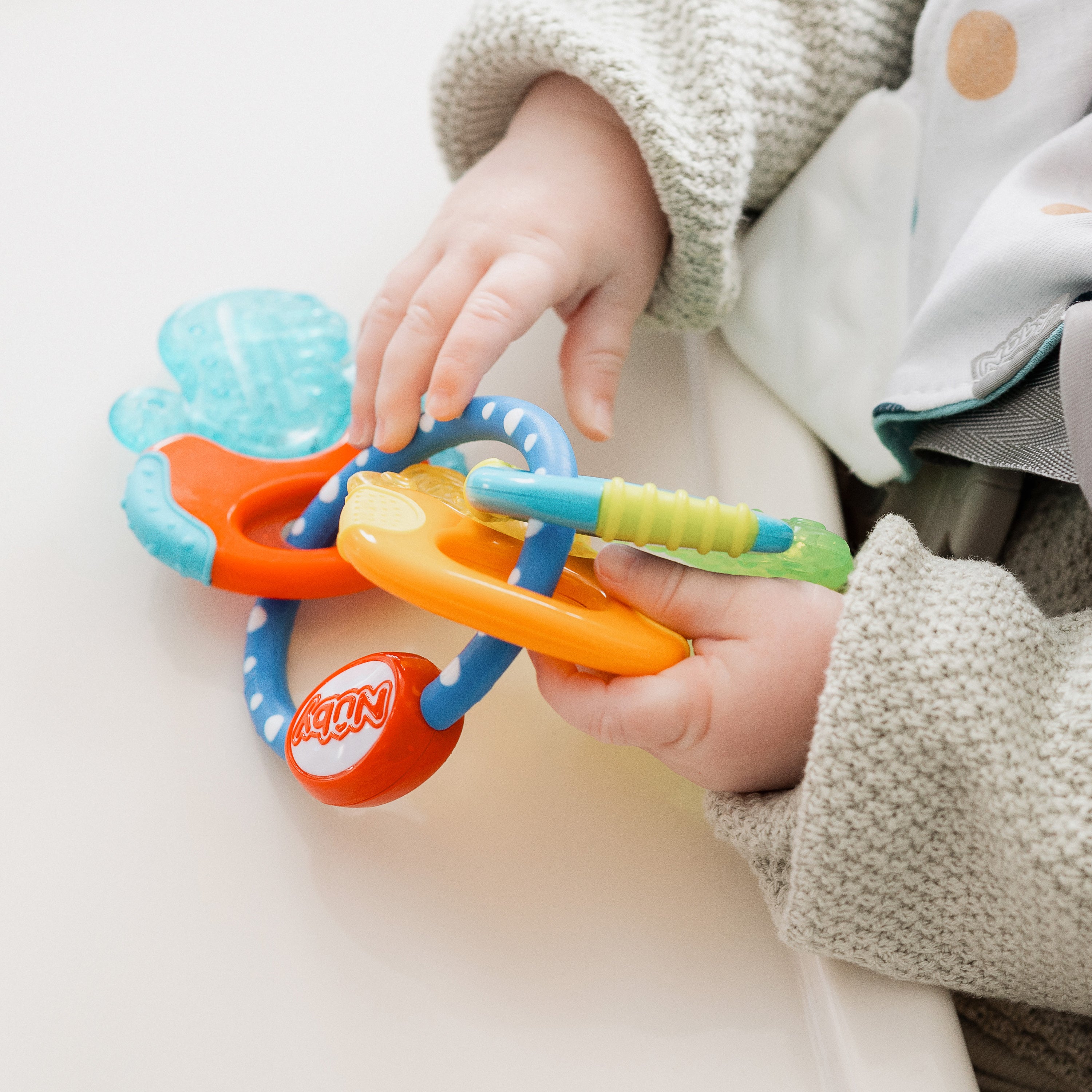 A baby in a light sweater holds IcyBite Teether Keys | Classic, a colorful teether featuring blue, orange, green, and yellow keys with various shapes and textures.