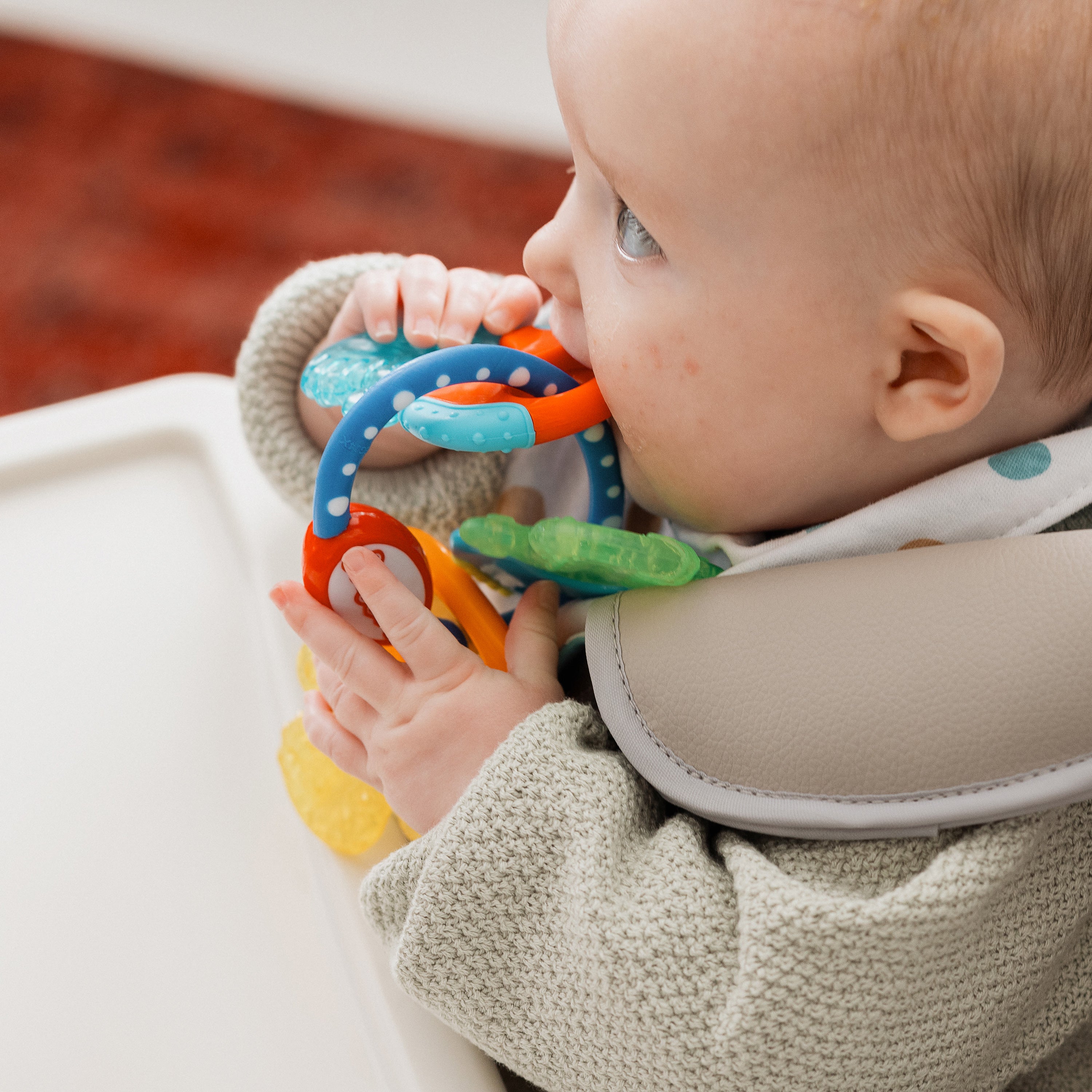 A baby in a high chair, wearing a grey sweater and a multicolored spotted bib, holds and chews on colorful IcyBite Teether Keys | Classic.