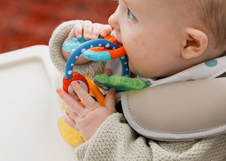 A baby in a high chair chews on IcyBite Teether Keys | Classic, holding them with both hands. Dressed in a light sweater and pastel dot bib, the baby enjoys soothing teething relief.