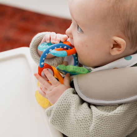 A baby in a high chair, wearing a grey sweater and a multicolored spotted bib, holds and chews on colorful IcyBite Teether Keys | Classic.