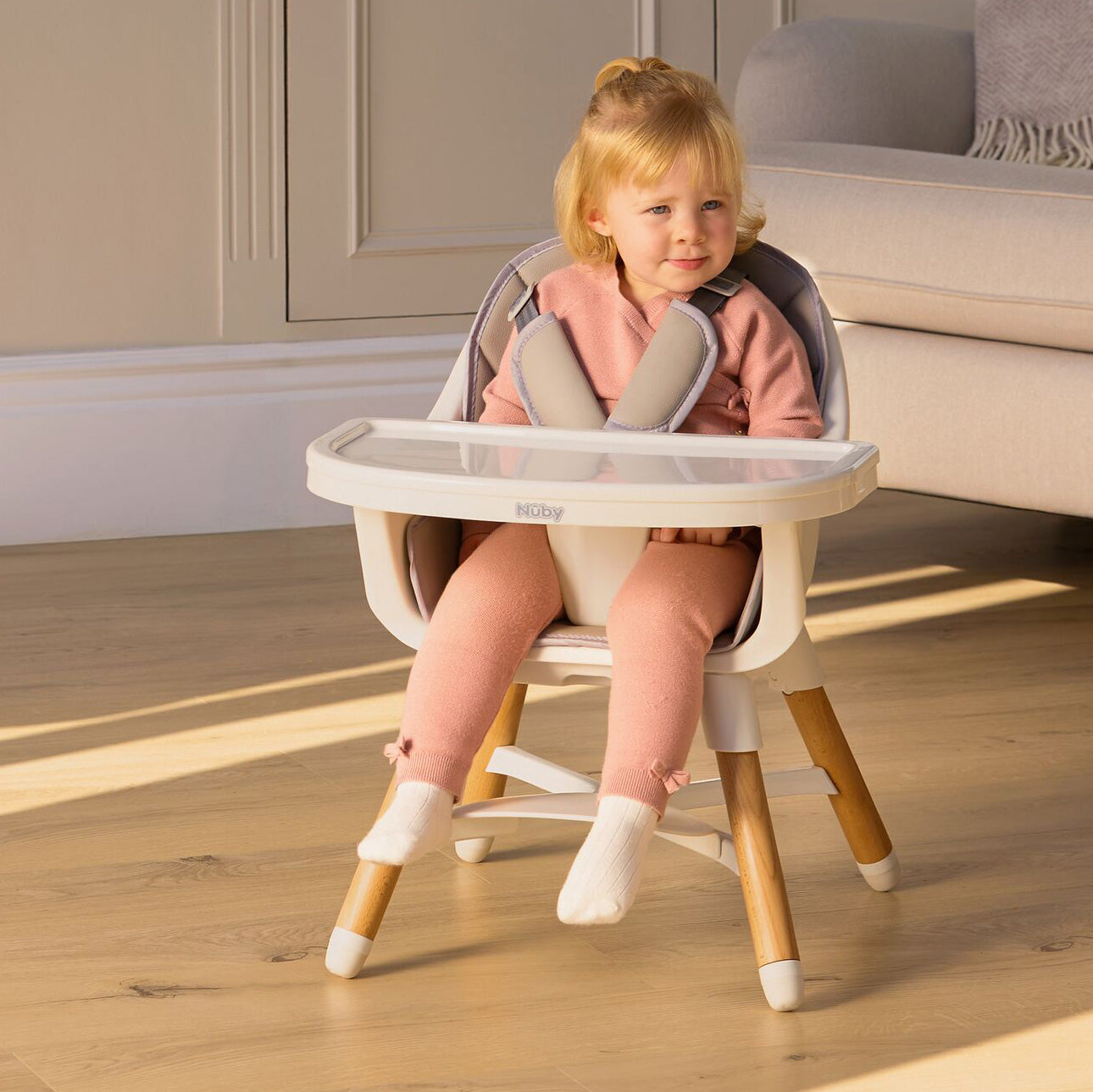A young child in a pink outfit and white socks sits in a Nuby 4-in-1 Multi-Use High Chair in Dark Beige, in a bright living room with wooden floors.