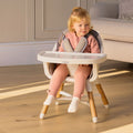 A young child in a pink outfit and white socks sits in a Nuby 4-in-1 Multi-Use High Chair in Dark Beige, in a bright living room with wooden floors.