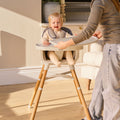 A smiling baby sits in the Nuby 4-in-1 Multi-Use High Chair in Dark Beige with a tray, as an adult adjusts it. The scene features light furniture and wooden floors in a bright, cozy room.