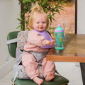 A smiling toddler with blond hair in a ponytail sits at a table in the Nuby Easy Go High Chair Booster Seat | Animals, wearing a pink outfit and purple bib, reaching for a green and blue sippy cup. A plant and window are in the background.