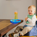 A young blonde toddler sits in a Nuby Easy Go High Chair Booster Seat | Bears, secured with adjustable straps. Wearing a green bib, he smiles at the table with a blue plate of food and a green sippy cup in front of him.