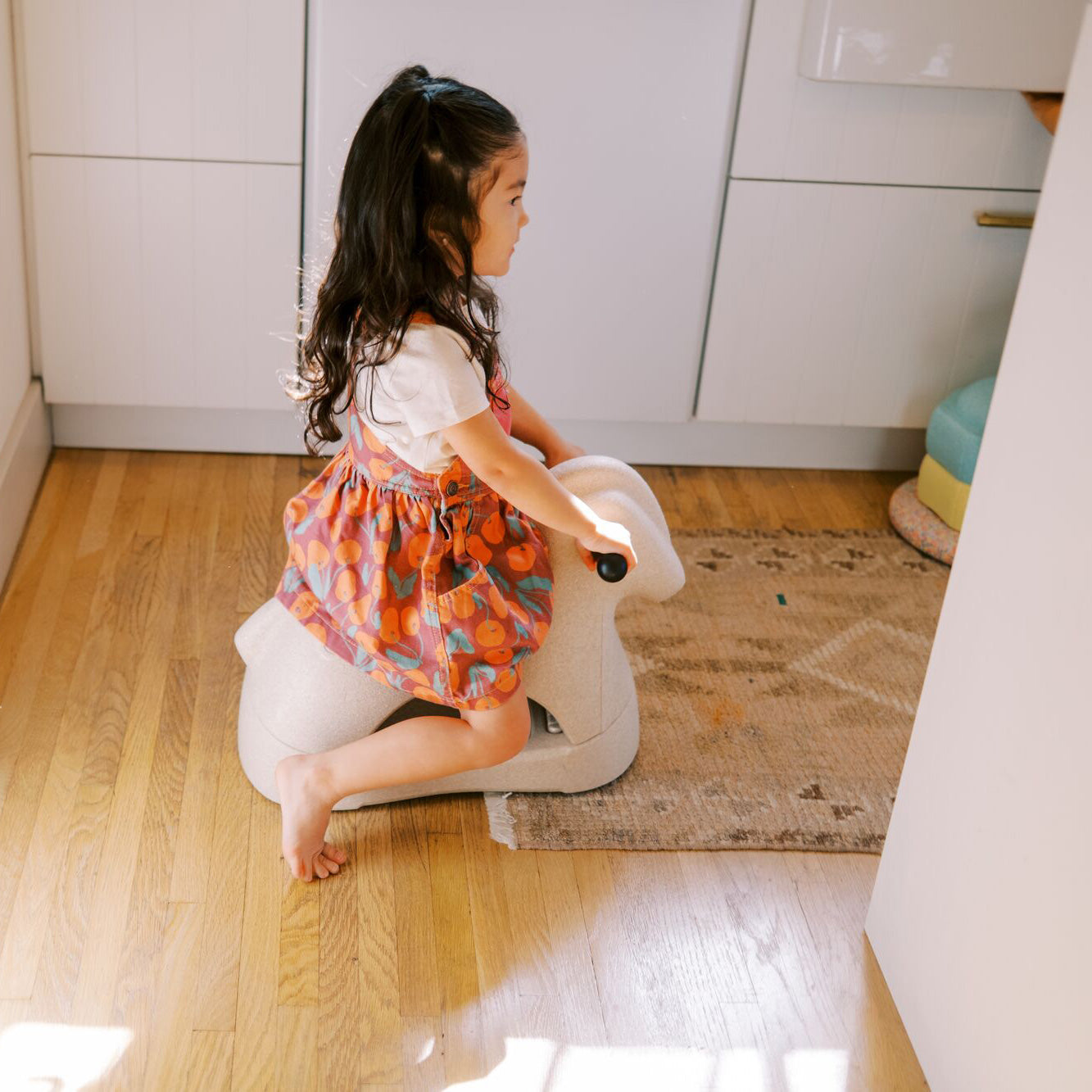 A young girl with long dark hair, in a colorful dress and white shirt, sits barefoot on the Rock 'N Ride | Beige in a bright kitchen with wooden floors and a textured rug.