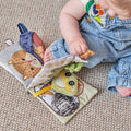 A baby in denim overalls sits on the floor, using one hand to turn a page of the Baby Sensory Book | City Bus—an engaging Montessori activity book with colorful city-themed illustrations and textures.