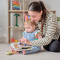 A woman sits on the floor with a toddler on her lap, guiding the child through a Baby Sensory Book | City Bus in a cozy room filled with books and toys.