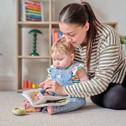 A woman sits on the floor with a toddler on her lap, guiding the child through a Baby Sensory Book | City Bus in a cozy room filled with books and toys.