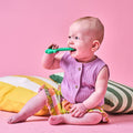 A baby in a purple top and colorful skirt sits on a pink floor, holding and chewing on a green Tool Teethers (4 Pack) silicone teether. Cushions with yellow and green stripes are behind the baby, set against a pink background.