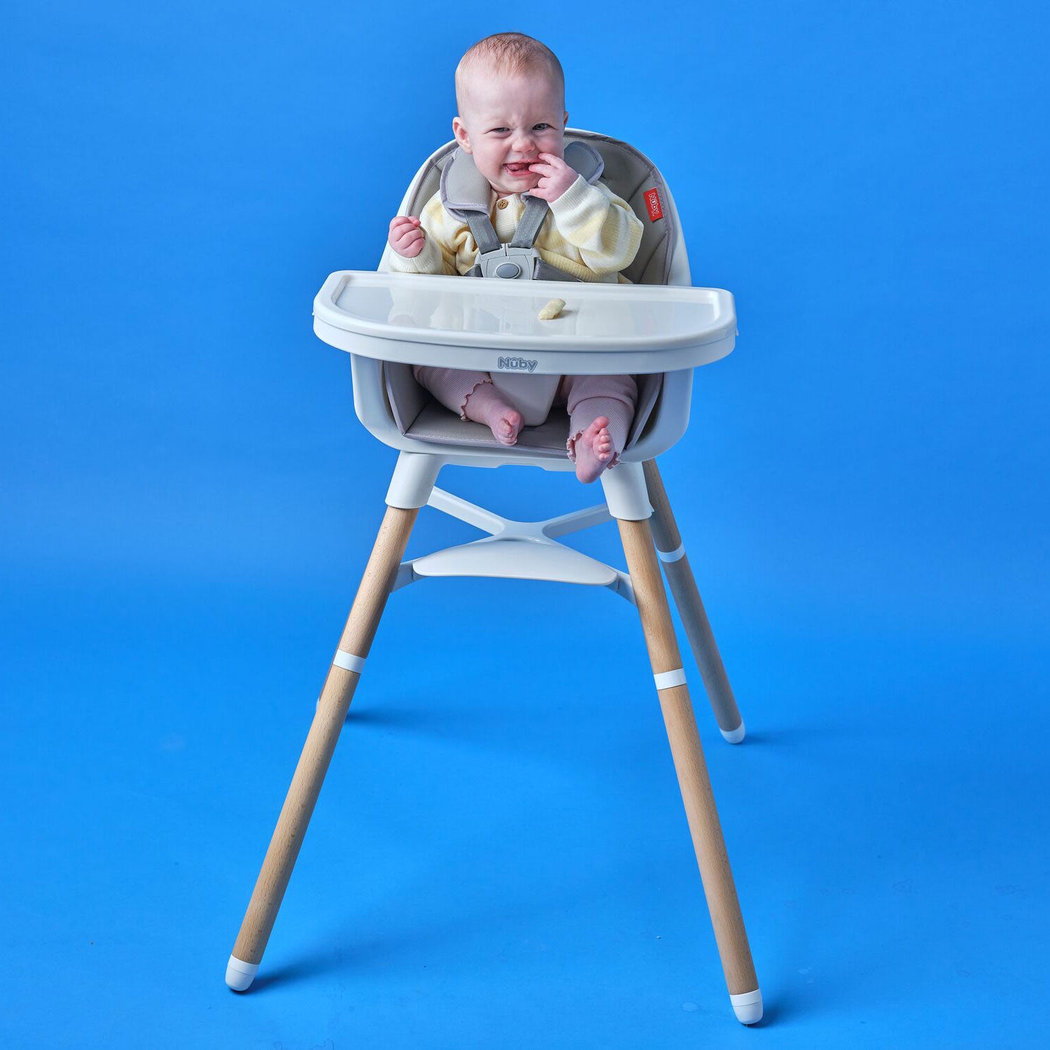 A baby in a light yellow outfit sits in the 4-in-1 Multi-Use High Chair | Dark Beige with a white tray, looking upset against a solid blue background. A few food pieces are scattered on the tray.