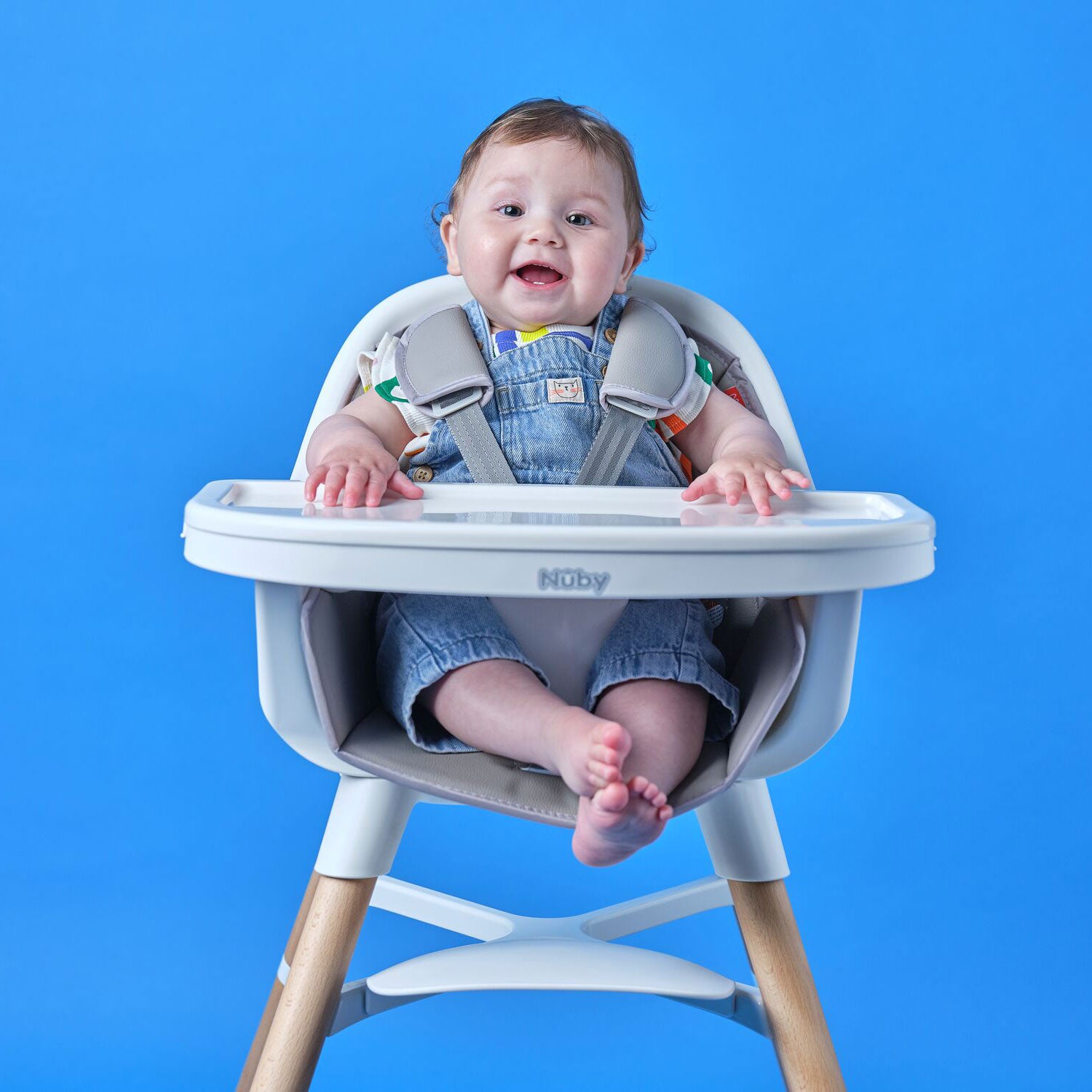 A smiling brown-haired baby with bare feet sits in the 4-in-1 Multi-Use High Chair | Dark Beige, hands resting on the tray, against a solid blue background. The baby wears denim overalls and looks happy in the versatile high chair.