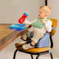 A young child smiles while sitting in a Nuby Easy Go High Chair Booster Seat | Bears at the table, wearing a green bib and holding a red sippy cup. In front of him is a blue divided plate filled with food.
