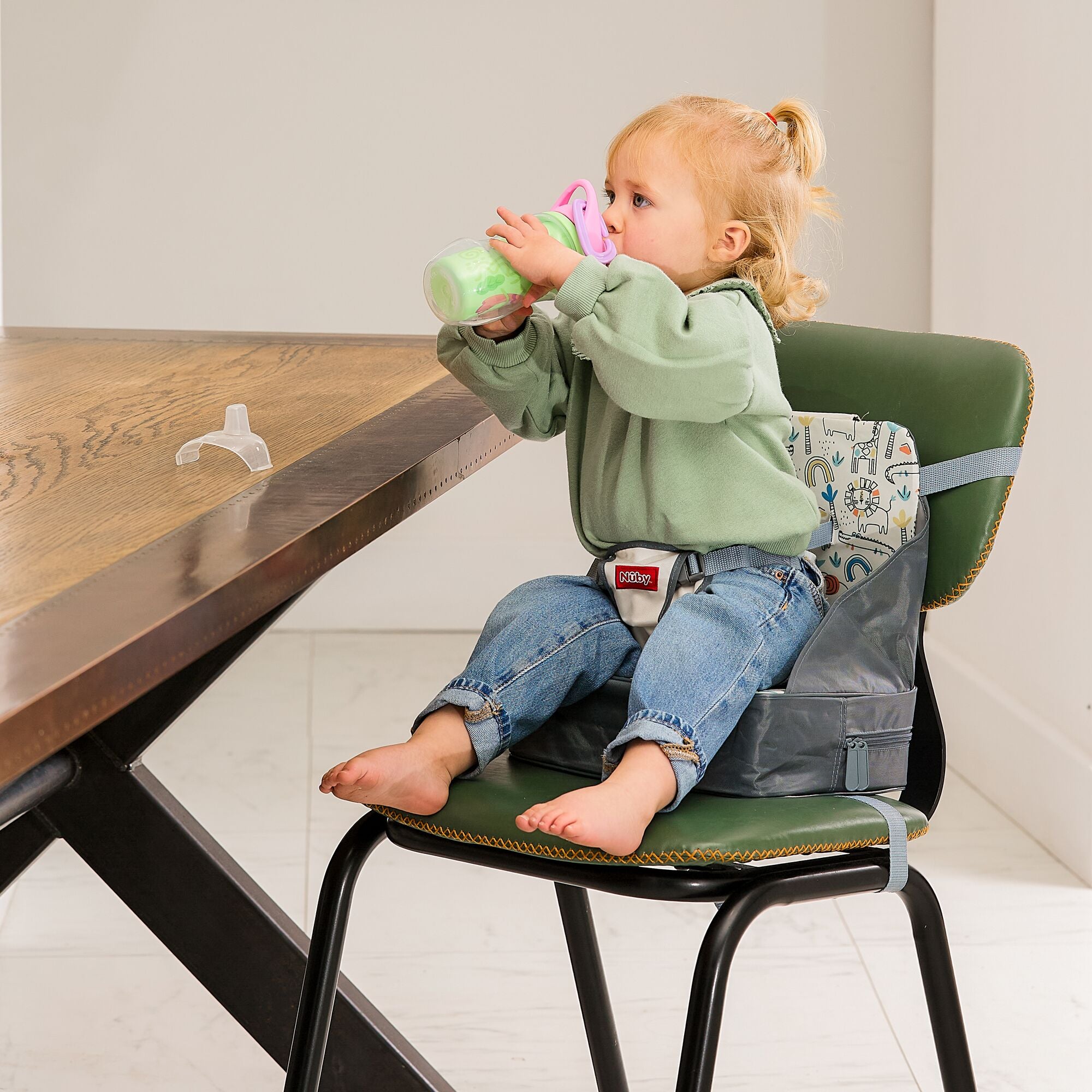 A toddler in a green sweater and jeans sits on the Nuby Easy Go High Chair Booster Seat | Animals at a wooden table, sipping from a cup. A pacifier rests on the table nearby.