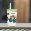 The Thirsty Kids Flexstraw Stainless Steel Cup | Monster Trucks features a no-spill straw and fun cartoon monster trucks, checkered flags, and stars. The Nûby logo in red appears on the cup, set outdoors.