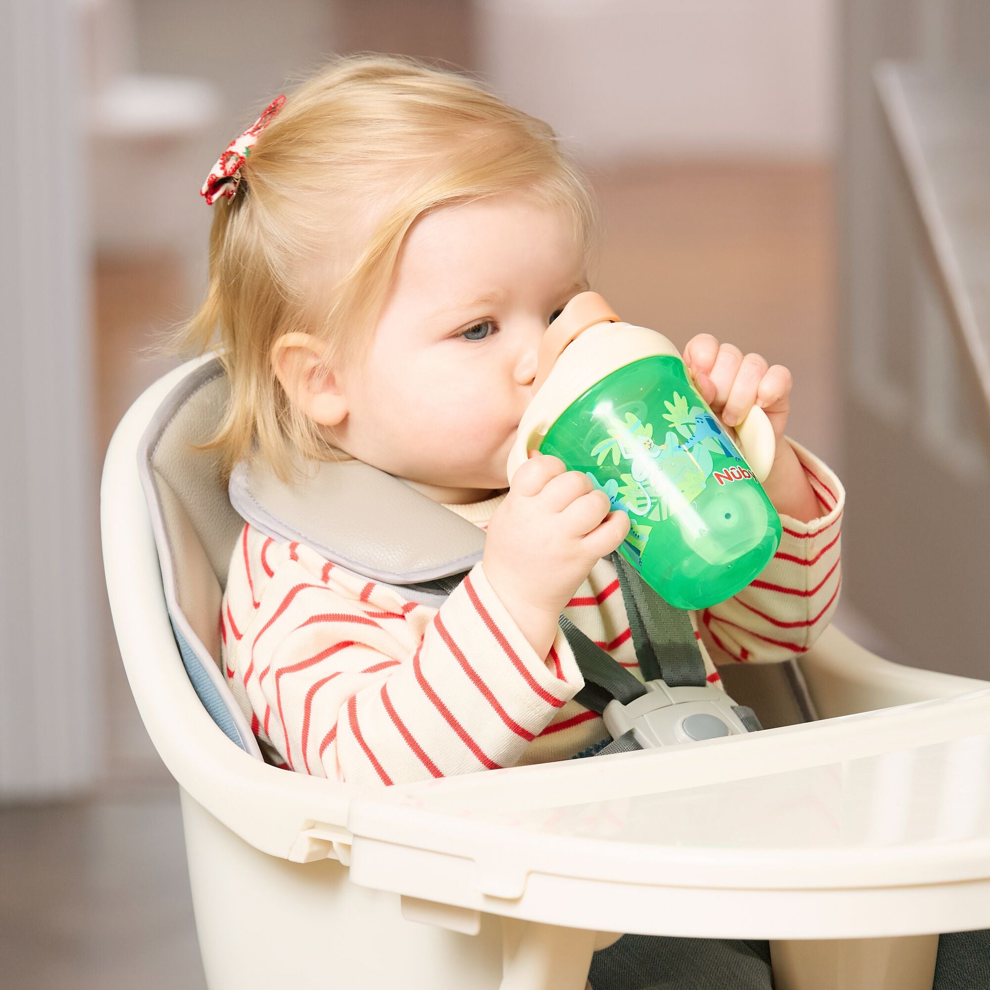 A young blonde child in a striped shirt sits in a high chair, drinking from the 2-in-1 Toddler Cup (2 Pack) | Jungle & Ocean, which features a weighted straw.