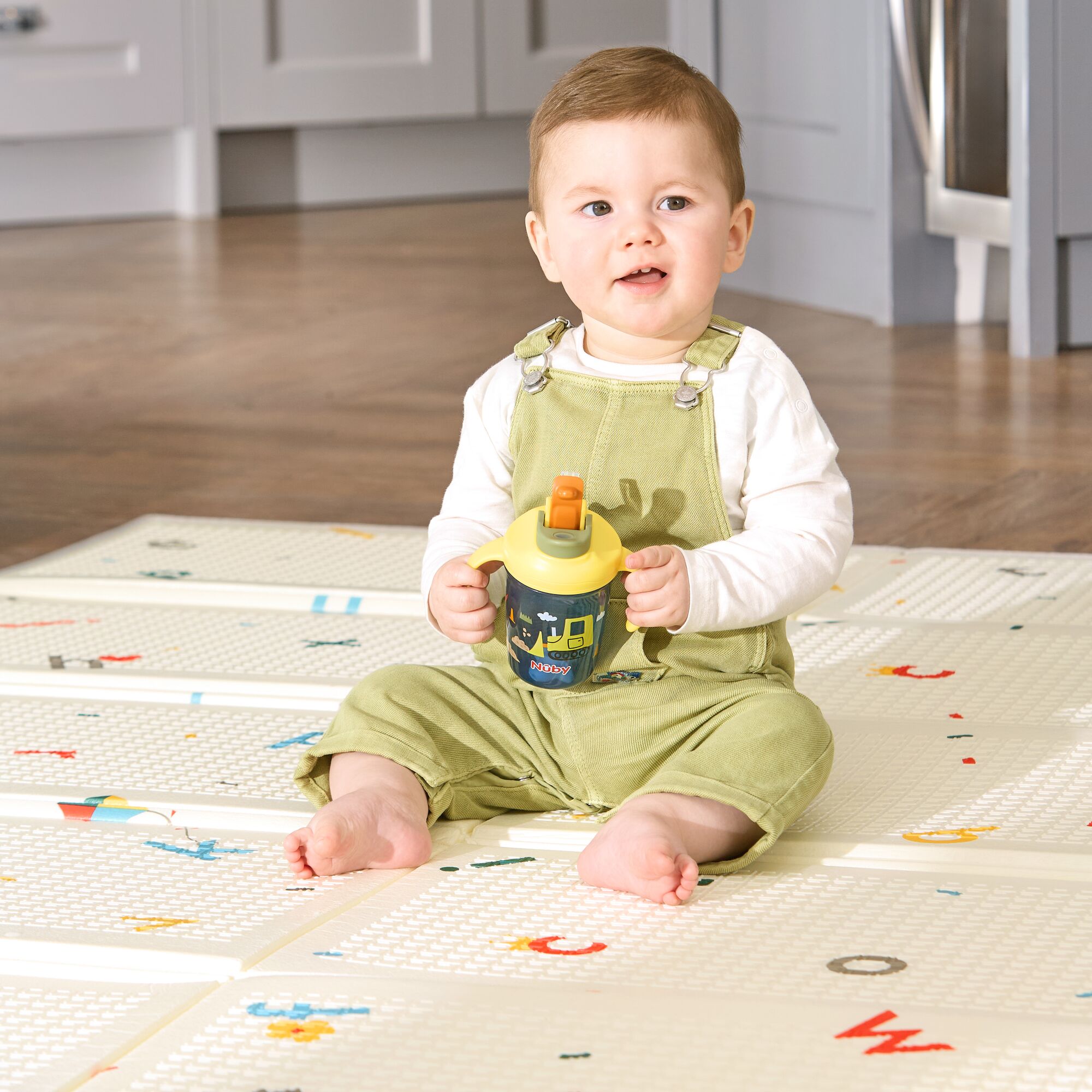 A baby in light green overalls and a white shirt sits barefoot on a patterned play mat, holding the 2-in-1 Toddler Cup (2 Pack) | Tractors & Monsters, with a kitchen visible in the background.