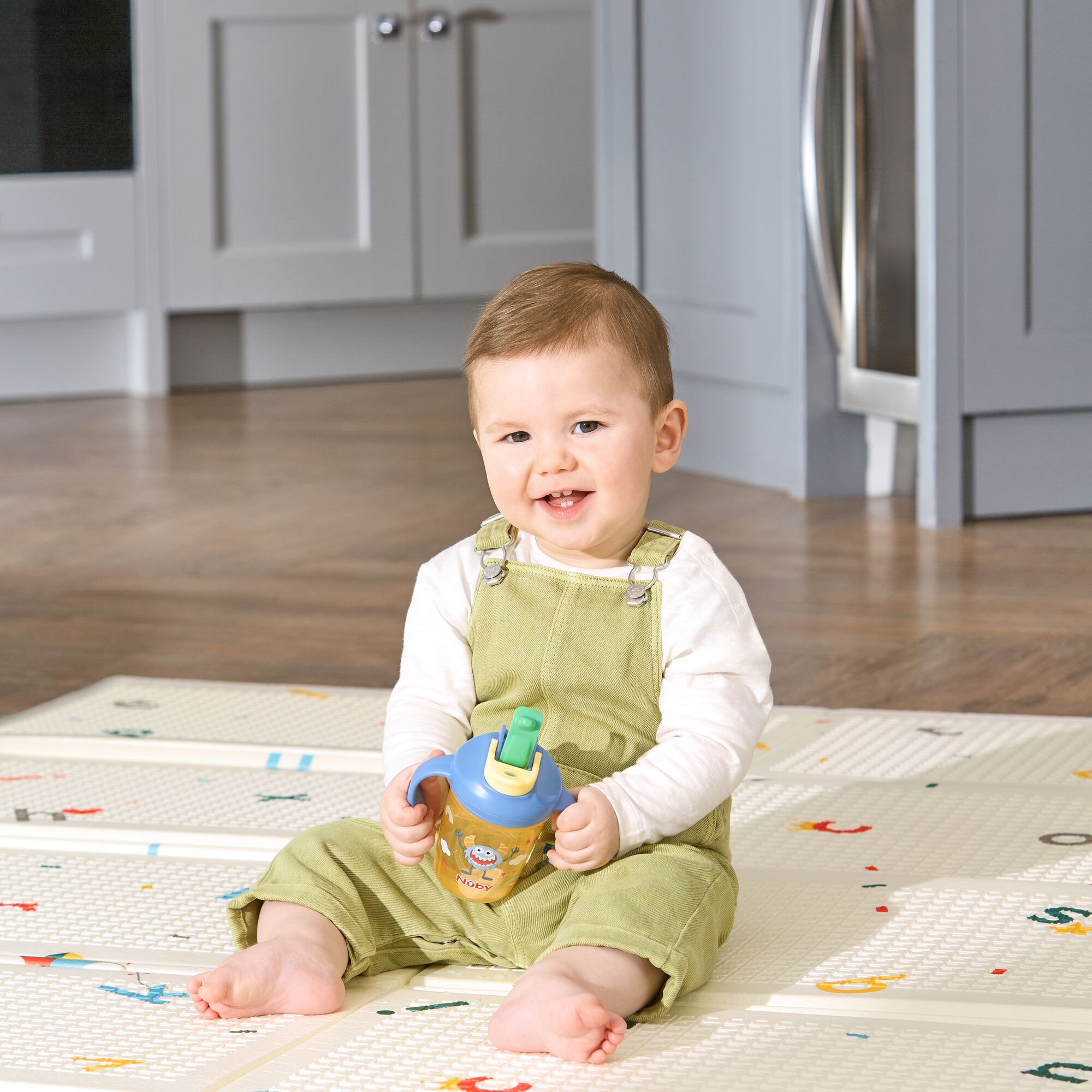 A smiling baby in light green overalls and a white shirt sits on a play mat, holding the 2-in-1 Toddler Cup (2 Pack) | Tractors & Monsters, with a kitchen in the background.