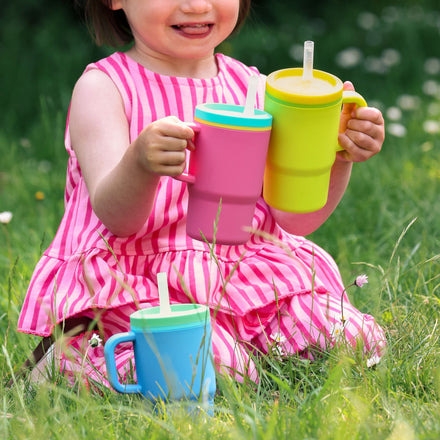 A child in a pink and white striped dress sits on the grass, smiling as she holds three colorful Silicone Tiny Tumbler | Blossom cups—blue, pink, and yellow—each topped with a white straw. Small white flowers are scattered around her.