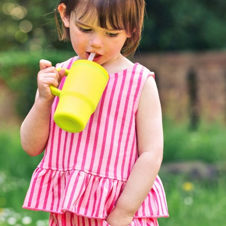 A young child in a pink and white striped outfit drinks from the Silicone Tiny Tumbler | Lemon Lime outdoors on a sunny day, with green grass and blurred plants in the background.