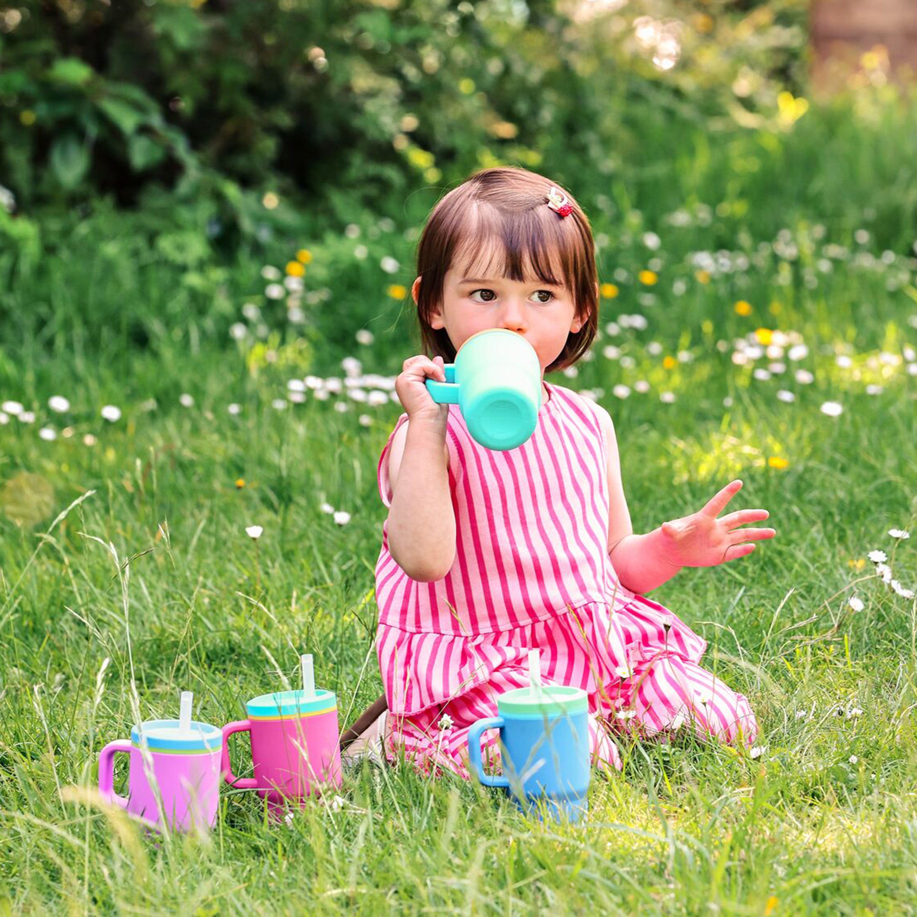 A young child in a pink striped dress sits on the grass, sipping from a green spill-proof cup with a silicone straw. Nearby, two colorful toddler tumblers are placed alongside the Silicone Tiny Tumbler in Lavender. Flowers and greenery fill the background.