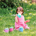 A young child in a pink striped dress sits on the grass, sipping from a green spill-proof cup with a silicone straw. Nearby, two colorful toddler tumblers are placed alongside the Silicone Tiny Tumbler in Lavender. Flowers and greenery fill the background.