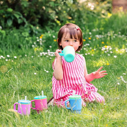 A young child in a pink striped dress sits on the grass, sipping from a green spill-proof cup with a silicone straw. Nearby, two colorful toddler tumblers are placed alongside the Silicone Tiny Tumbler in Lavender. Flowers and greenery fill the background.