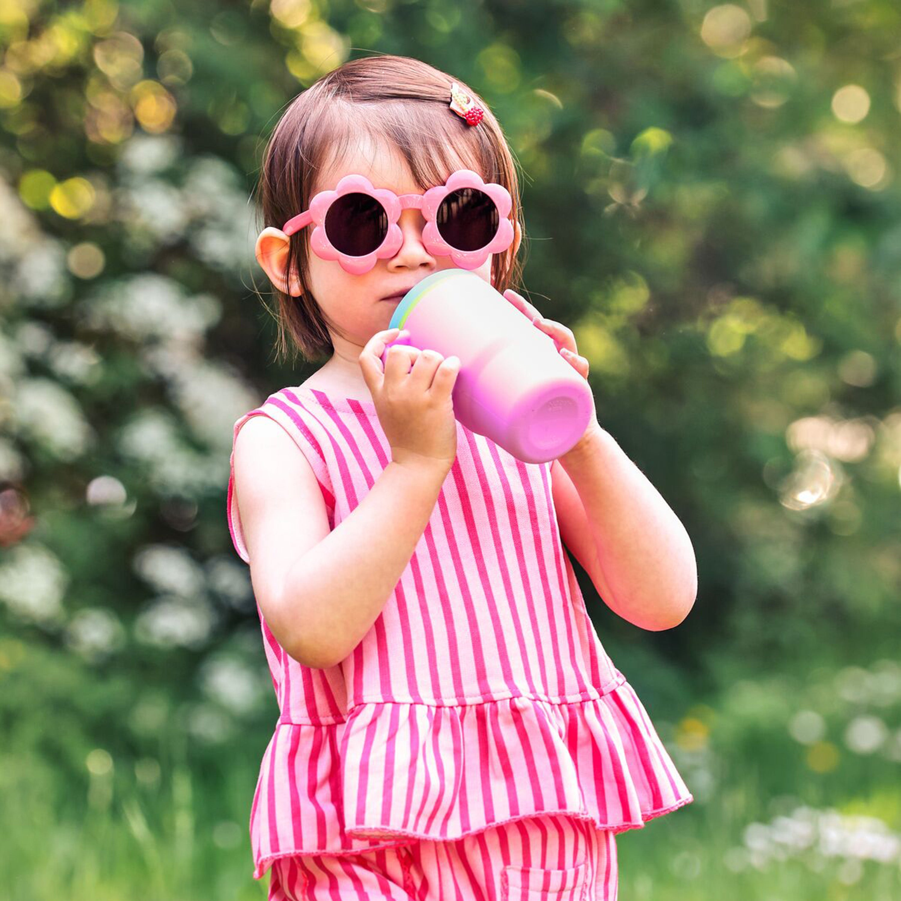 A young child wearing pink sunglasses and a pink striped outfit drinks from the Silicone Tiny Tumbler | Blossom outdoors, surrounded by greenery and blurred flowers.