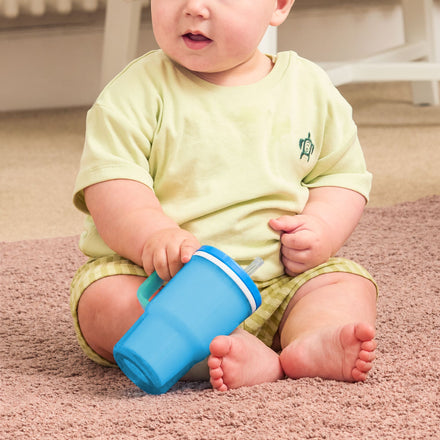 A baby in a yellow outfit sits on a carpeted floor, holding the Insulated Tiny Tumbler in Electric Blue.