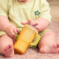 A baby dressed in light green clothing, with a shirt featuring a small turtle design, sits on the carpet holding the Insulated Tiny Tumbler | Orange Crush—a no-spill sippy cup with a silicone straw. Only the baby's lower face, arms, and legs are visible.
