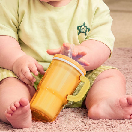A baby dressed in light green clothing, with a shirt featuring a small turtle design, sits on the carpet holding the Insulated Tiny Tumbler | Orange Crush—a no-spill sippy cup with a silicone straw. Only the baby's lower face, arms, and legs are visible.