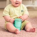 A baby in a yellow outfit sits on a pink carpet, holding the Insulated Tiny Tumbler | Seafoam with a silicone straw sippy cup. Only the baby's bottom half of the face and body are visible.