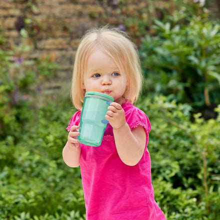 A young child with blonde hair in a pink shirt drinks from the Insulated Tiny Tumbler in Seafoam outdoors, surrounded by green foliage and a stone wall.