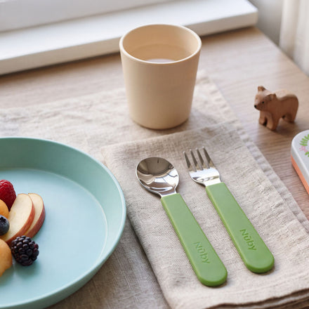 A plate with apple slices and berries sits on a wooden table near a window, alongside the Stainless Steel Toddler Utensil Set | Forest Animals—featuring green-handled spoon and fork—resting on a napkin, a beige cup with a drink, and a small wooden animal figurine.