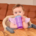A baby with light brown hair sits in a high chair, holding and drinking from the Insulated Tiny Tumbler in Lavender. There is a wooden table in front and a brown couch in the background.