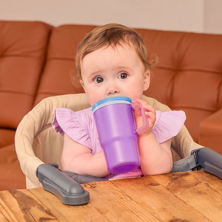 A baby with light brown hair sits in a high chair, holding and drinking from the Insulated Tiny Tumbler in Lavender. There is a wooden table in front and a brown couch in the background.