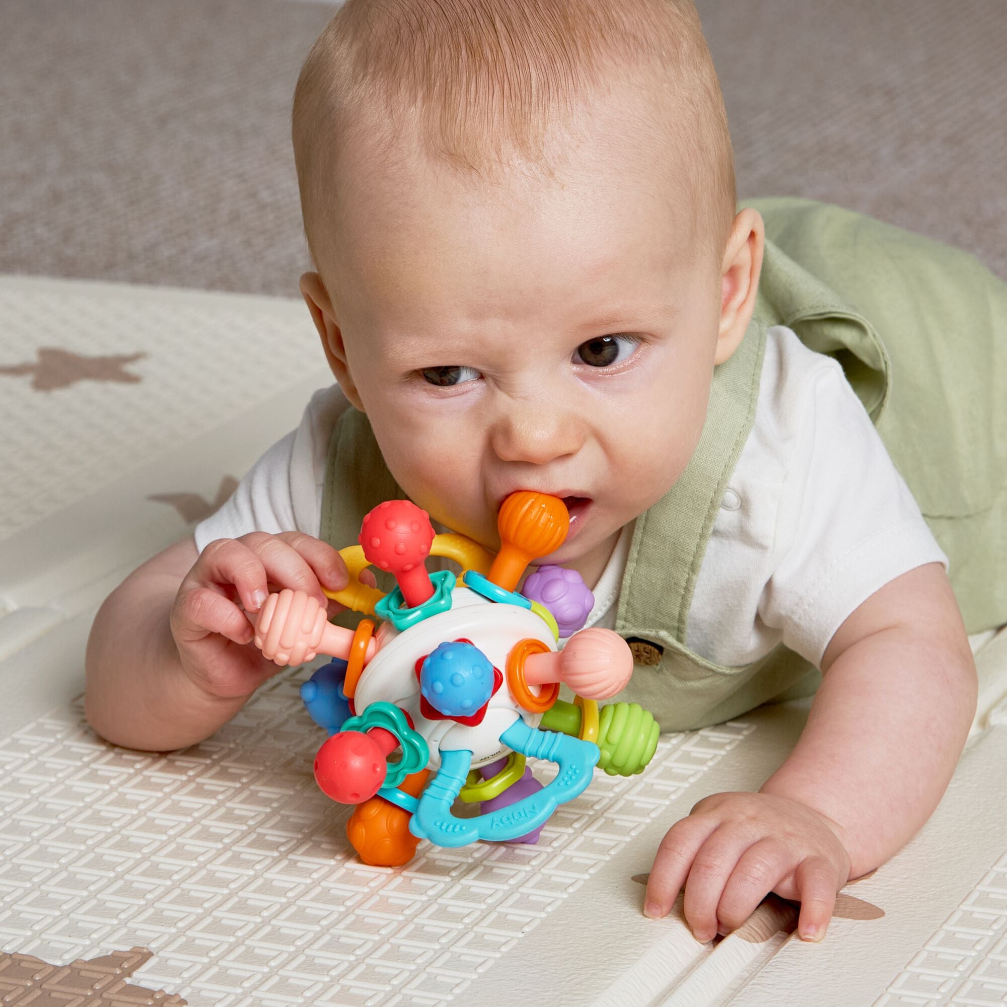A baby in a white shirt and light green overalls lies on a play mat, holding and chewing on the Twist Ball Teether for teething relief.