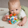 A baby in a white shirt and light green overalls lies on a play mat, holding and chewing on the Twist Ball Teether for teething relief.