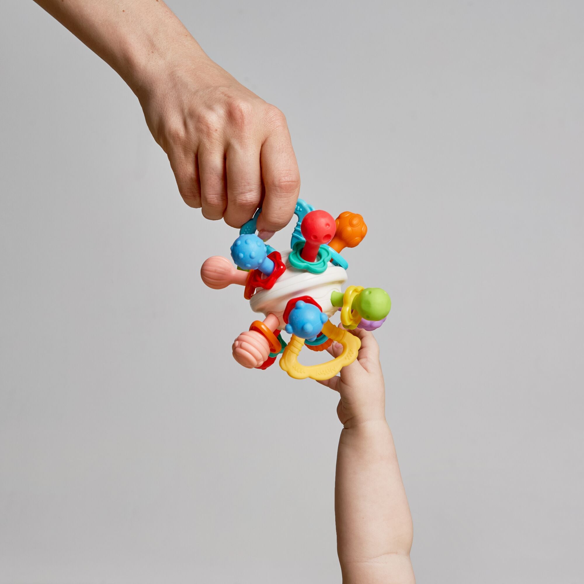 An adult hand passes the Twist Ball Teether—a colorful sensory toy—to a baby’s hand against a plain gray background.