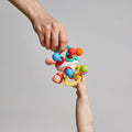 An adult hand passes the Twist Ball Teether—a colorful sensory toy—to a baby’s hand against a plain gray background.